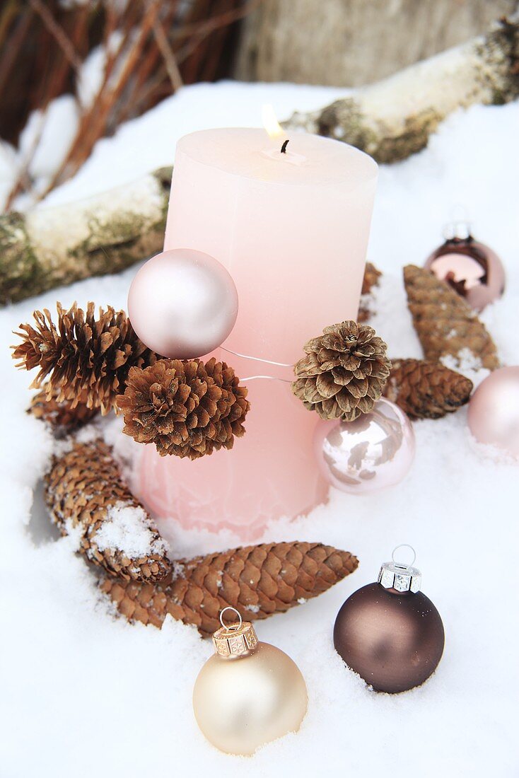 A pink candle with pine cones and Christmas baubles in the snow