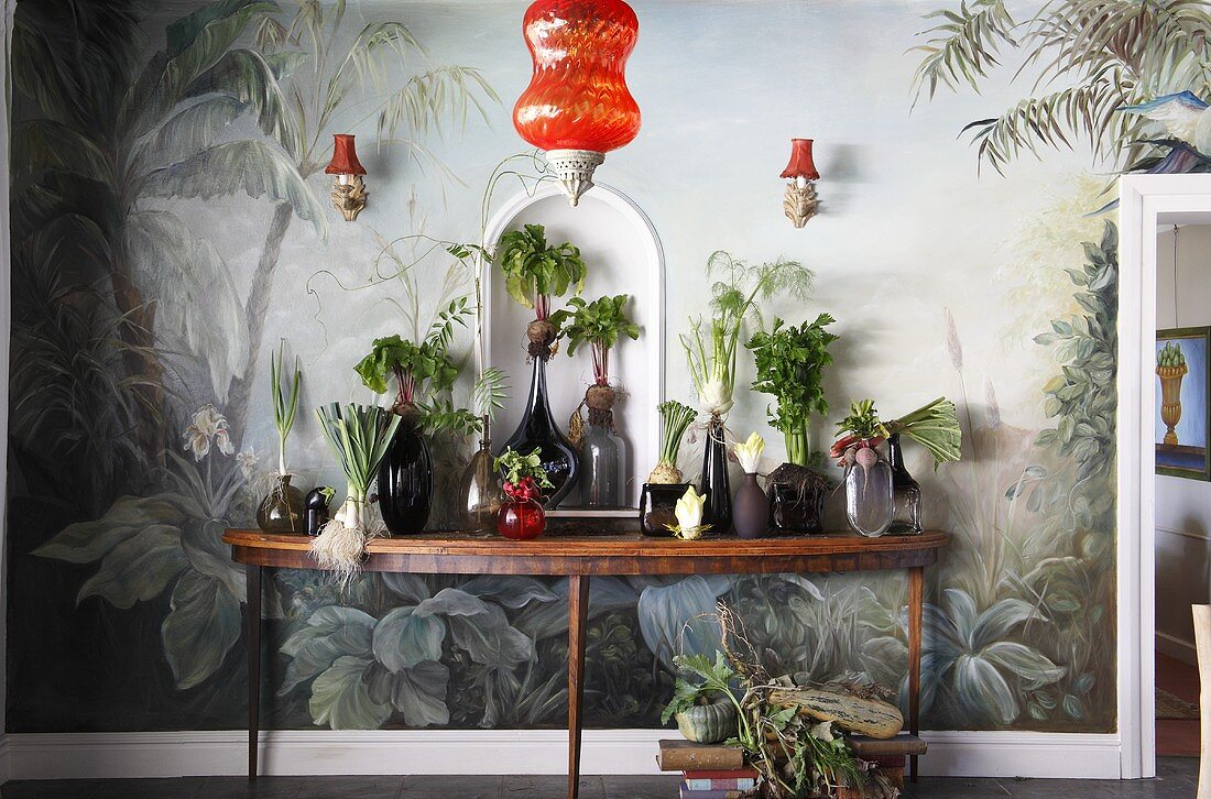 Assorted vegetables in glass vases on a console table in front of a painted wall