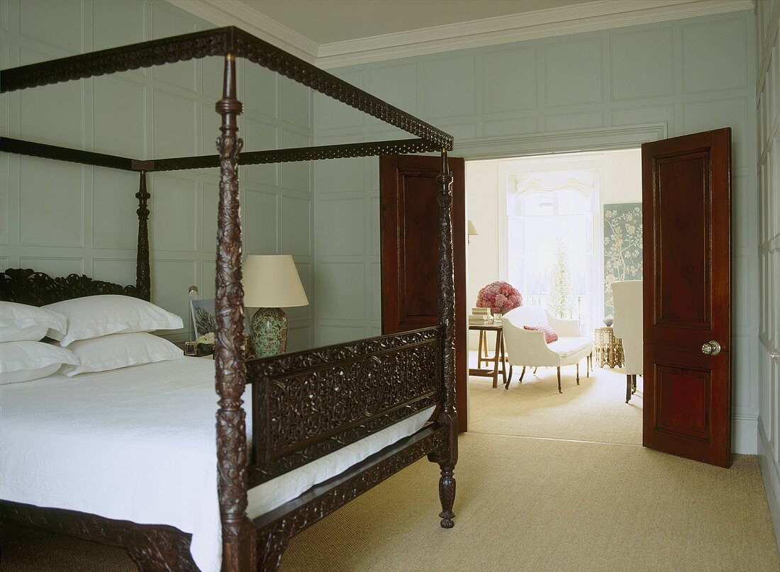 A traditional bedroom with painted panelling, carved wooden four poster bed, open double doors to sitting room