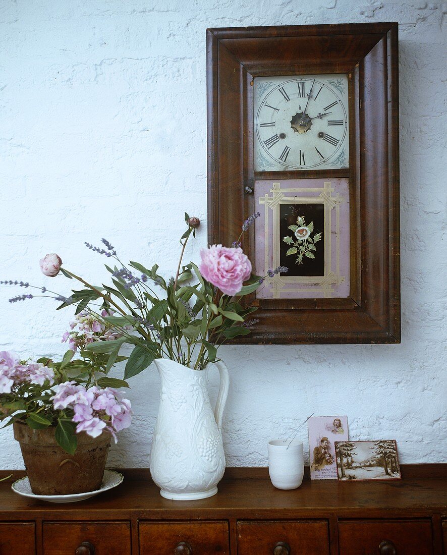 Flowers in a white jug on a wooden cupboard and a framed wall clock