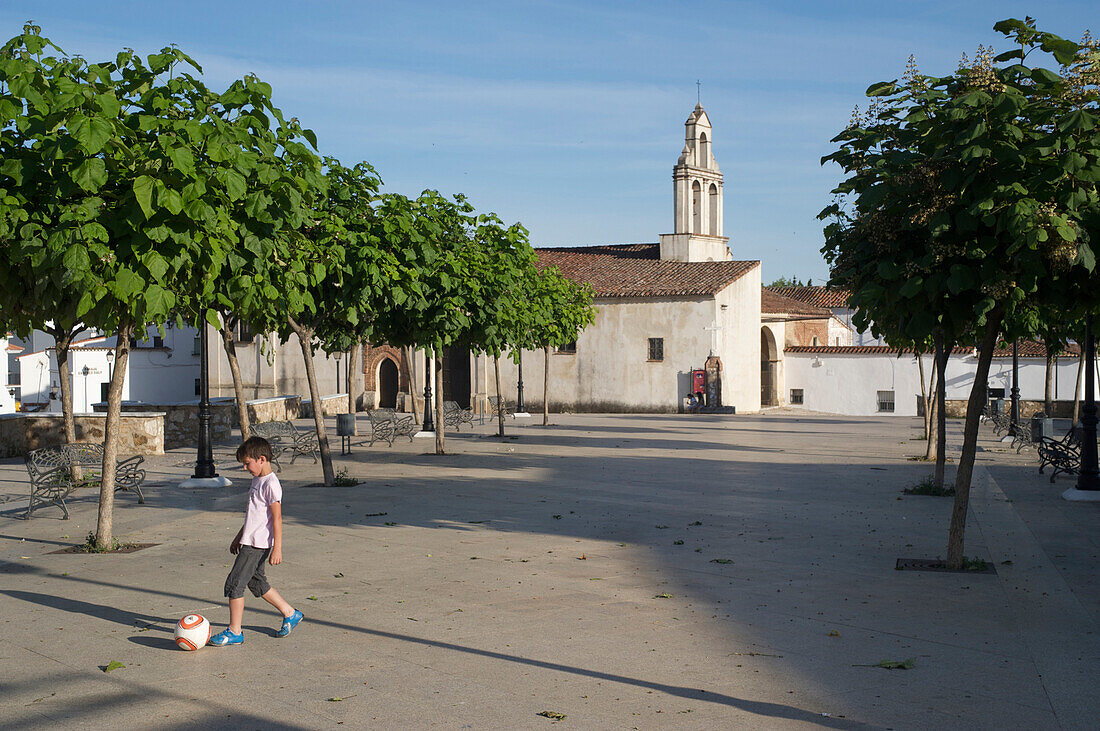 Junge mit Fussball auf Platz mit kleiner Kirche in der Altstadt von Arcena, Huelva, Andalusien, Spanien