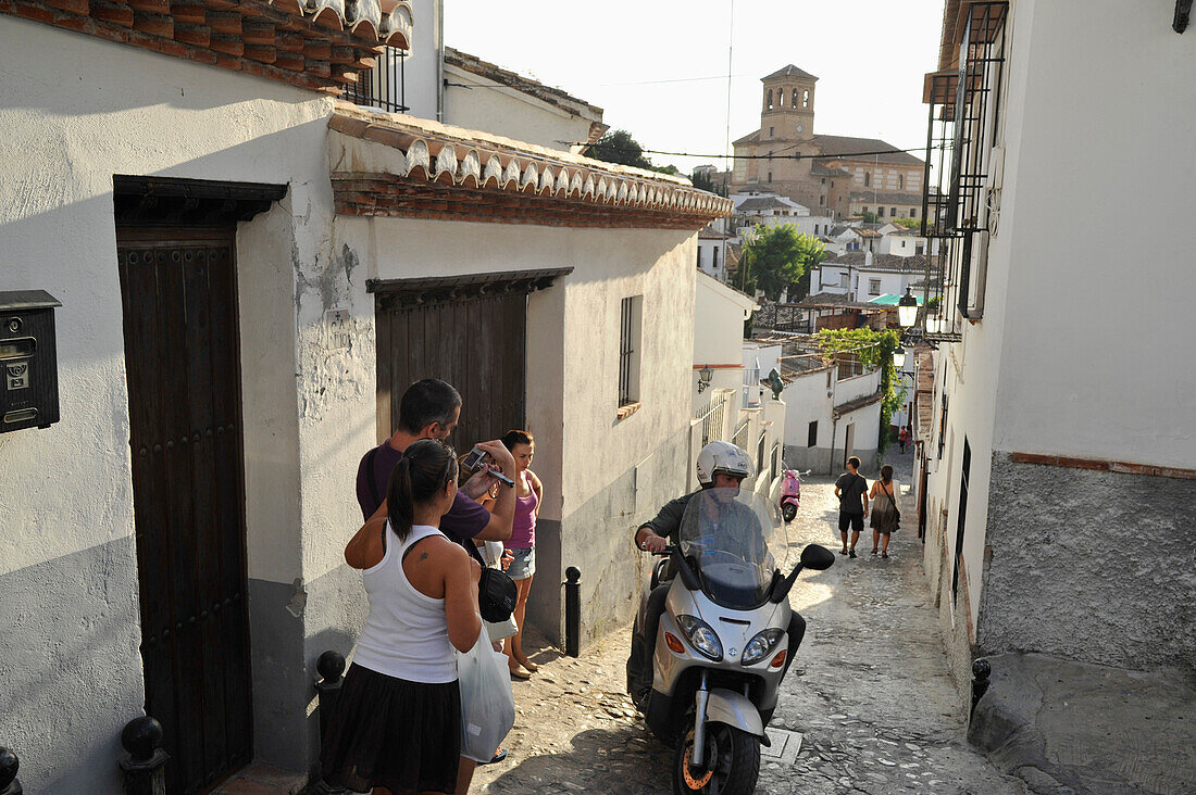 Menschen und Motorroller auf einem gepflasterten Weg im Sacromonte, Alhambra im Hintergrund Granada, Andalusien, Spanien