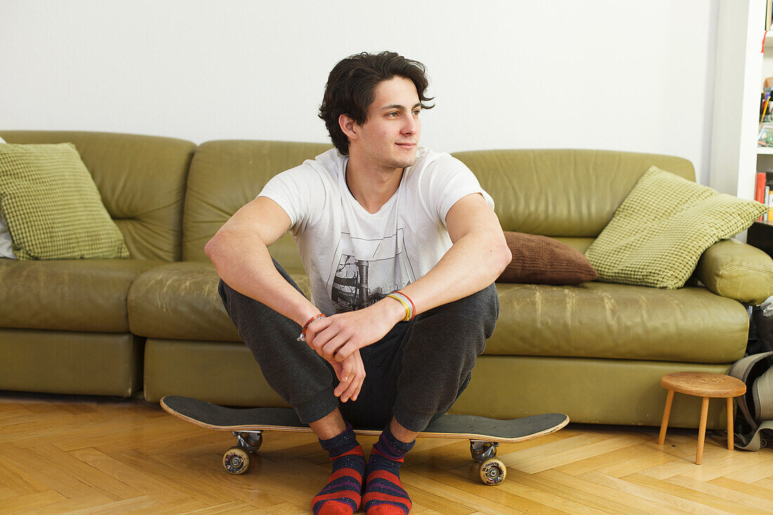 Young man at home, sitting on a skateboard, indoors
