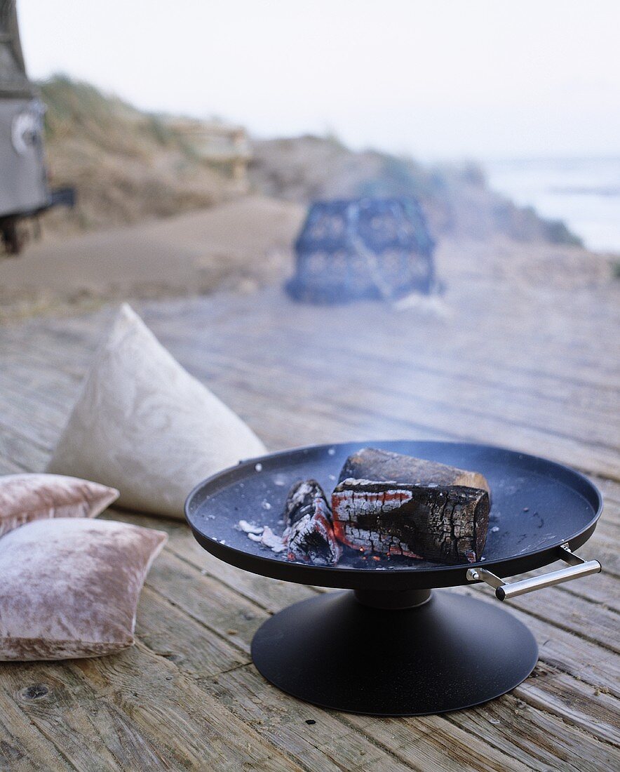 Holzscheiten mit Glut in Feuerschale auf Terrasse und Meerblick