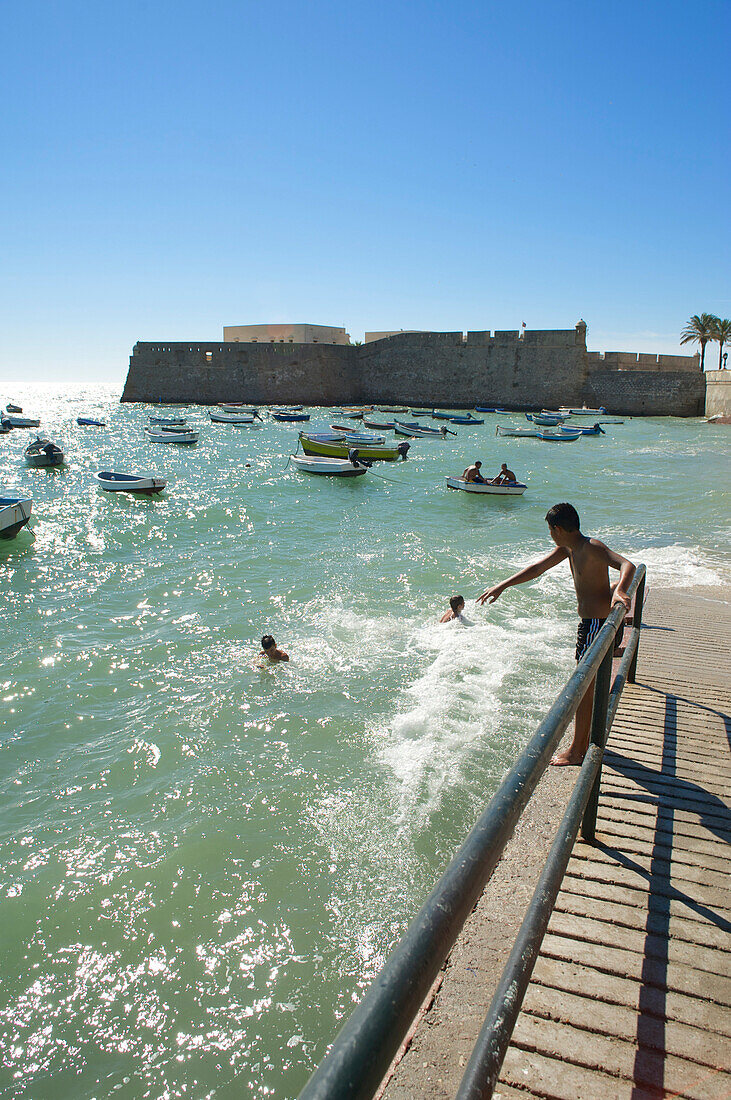 Boote und und Junge der von Mauer ins Meer springen will beim Castillo de Santa Catalina, Strand von Cadiz, Cadiz, Andalusien, Spanien