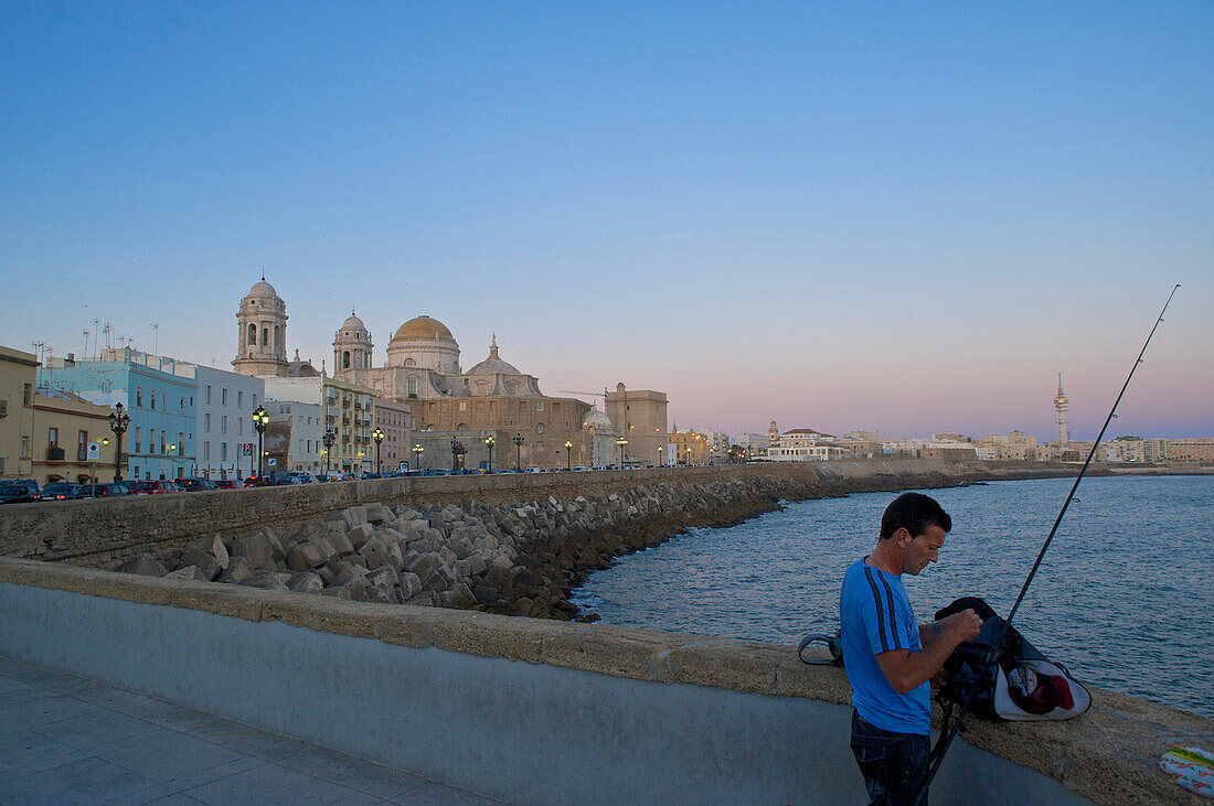 Man fishing from the promenade along the Av. Campo del Sur with wall and old style electric lights, view over the sea to the cathdral, Cadiz, Andalusia, Spain, Europe