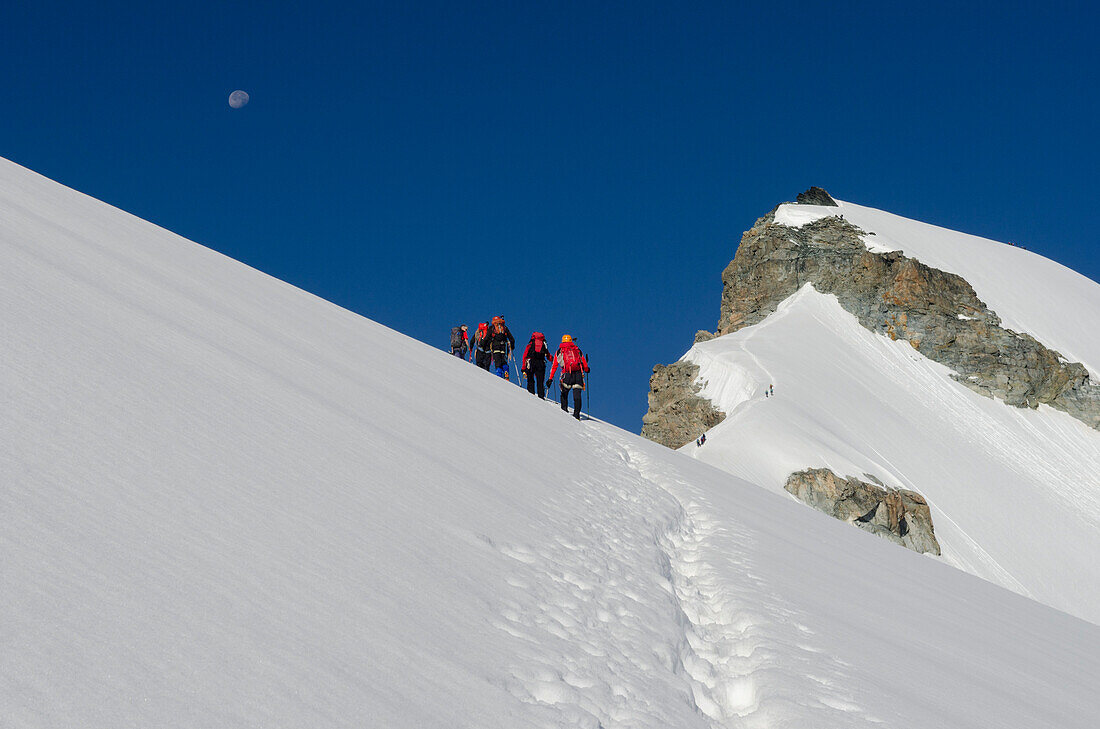 A group of alpinists on the snow crest of Hohlaub Ridge ascending towards the summit of Allalinhorn, above them the moon, Pennine Alps, canton of Valais, Switzerland