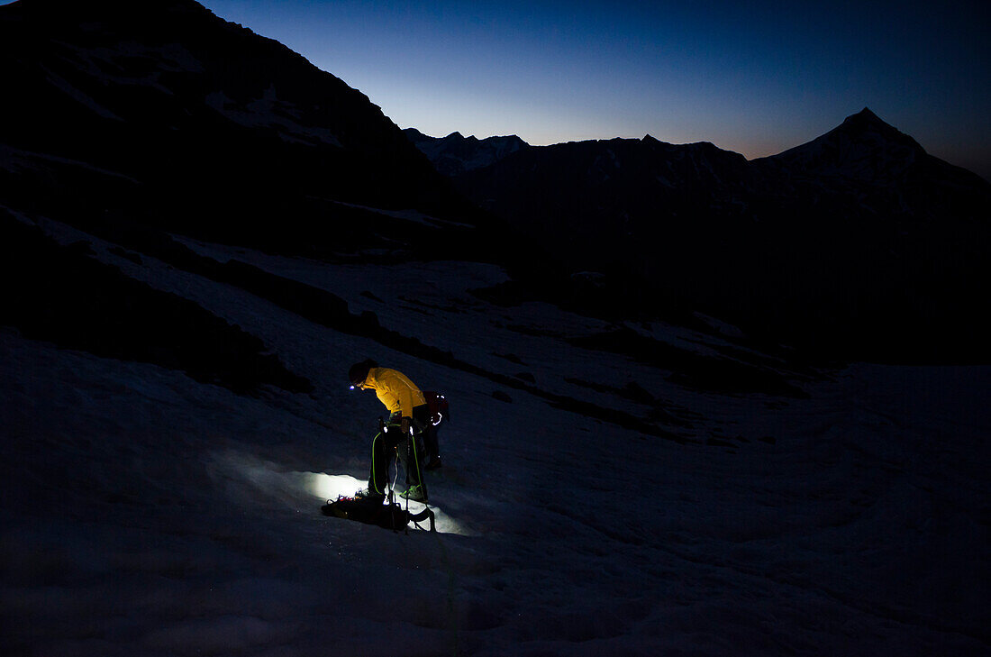 In the light of her headlamp, a female alpinist is putting on her crampons on the Hohlaub Glacier, in the background the first light of dawn, Pennine Alps, canton of Valais, Switzerland