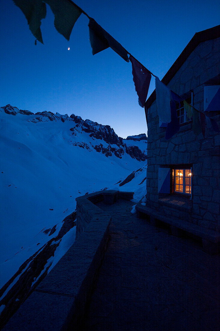 Im Stubenfenster der Bächlitalhütte brennt Licht, während es draußen dämmert und der Mond scheint, Bächlital, Grimselgebiet, Berner Alpen, Kanton Bern, Schweiz