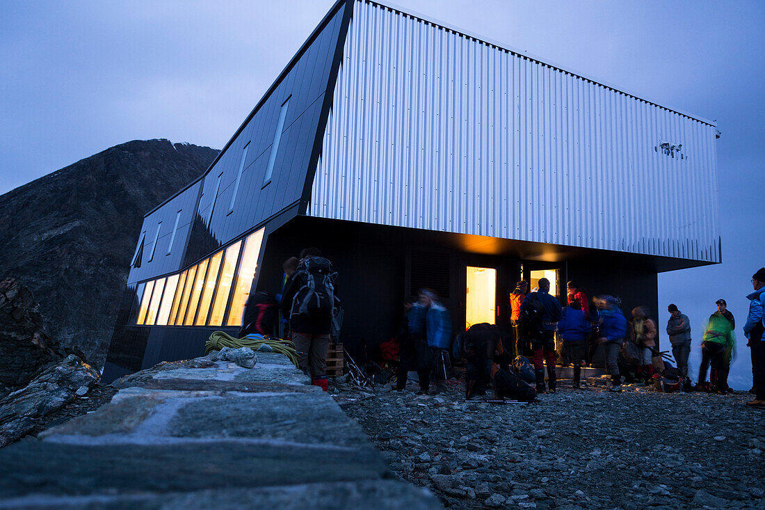 Früh morgens bereiten sich Gäste vor der neuen, modernen Hütte namens Cabane de Tracuit für die Tour aufs Bishorn vor, während die Fenster der Hütte in der Morgendämmerung leuchten, Val d‘Anniviers, Walliser Alpen, Kanton Wallis, Schweiz