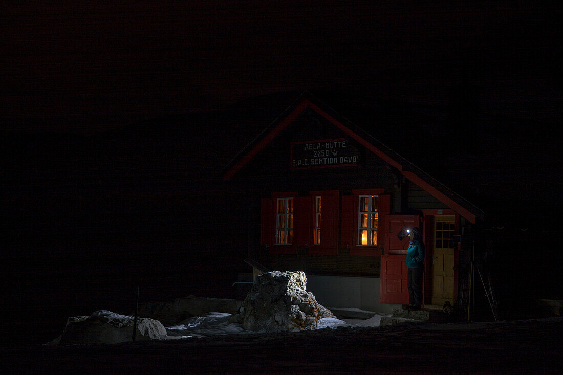 A female alpinist with her headlamp on standing in the dark in front of the door of the alpine hut called Chamonas d‘Ela, above the village of Berguen, Albula region, Grison Alps, canton of Grison, Switzerland