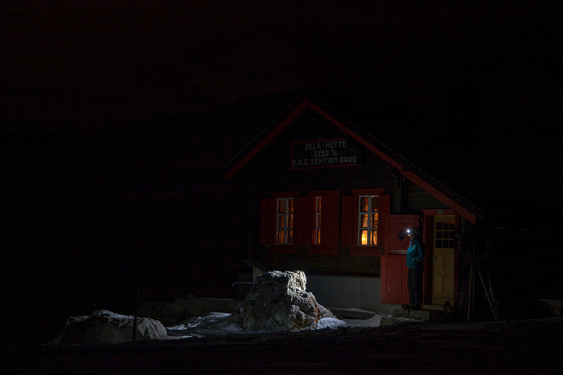 Eine Bergsteigerin und mit einer leuchtenden Stirnlampe steht nachts vor der Tür der Hütte namens Chamonas d‘Ela, oberhalb des Dorfs Bergün, Albulagebiet, Bündner Alpen, Kanton Graubünden, Schweiz