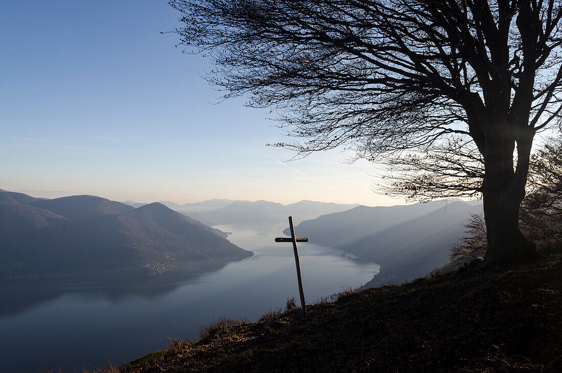 Ein Gipfelkreuz und ein Laubbaum auf dem Gipfel namens Corona dei Pinci, dahinter der Lago Maggiore und die Hügel diesseits und jenseits der italienisch-schweizerischen Landesgrenze, Kanton Tessin und Region Lombardei, Schweiz und Italien