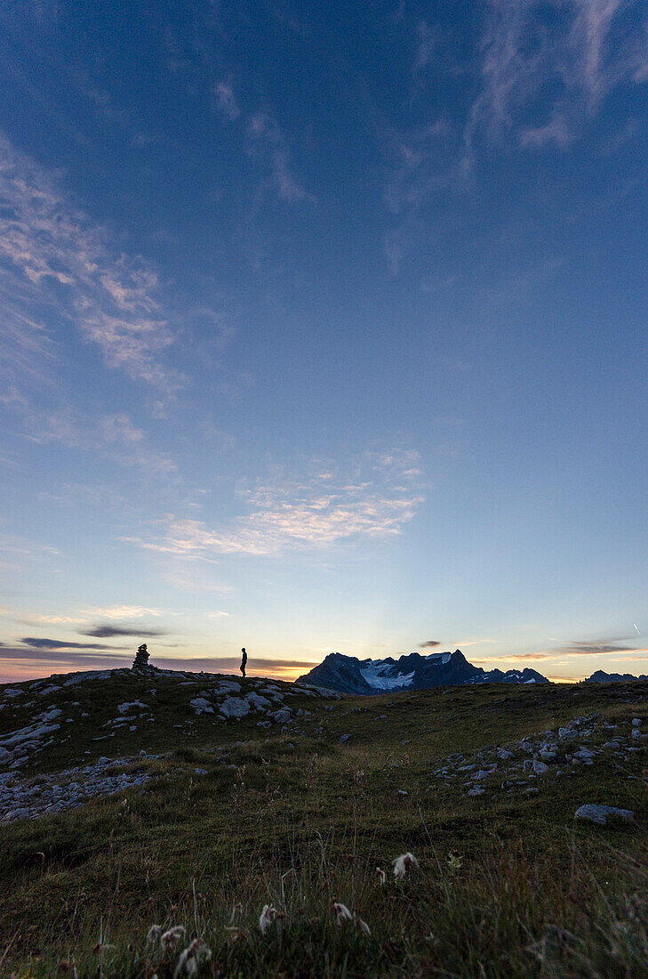 In the first light of the day a hiker is approaching a cairn on the carst mountain called Silberen, in the background the summits of the Glaernisch massif, Glarus Alps, cantons of Schwyz and Glarus, Switzerland