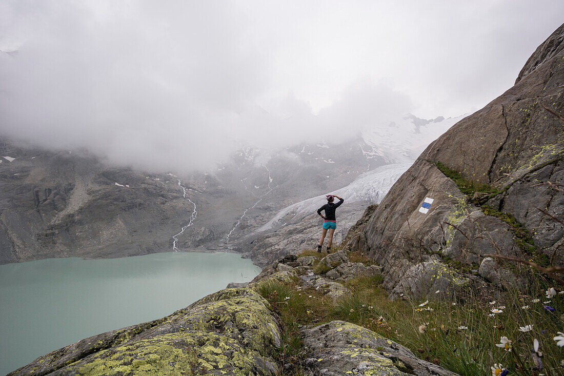 Eine junge Frau in Wanderausrüstung steht auf einem aussichtsreichen Alpinwanderweg und blickt auf den Nebel verhangenen Gauligletscher, darunter der Gaulisee, Gauligebiet, Berner Alpen, Kanton Bern, Schweiz