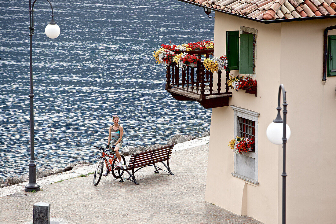 Junge Frau macht mit ihrem Fahrrad eine Pause an einer Bank bei einem See, Gardasee, Italien