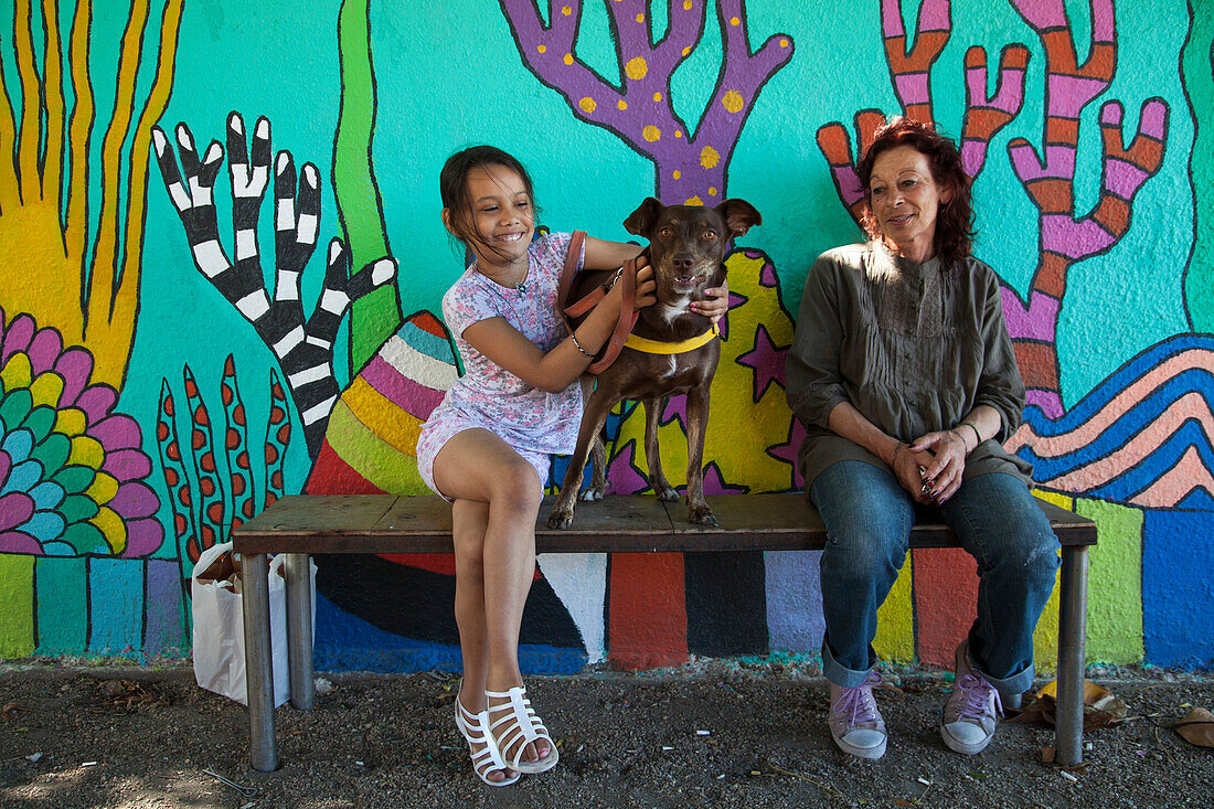 Daughter, dog and mother on bench in front of wall mural, Montevideo, Montevideo, Uruguay