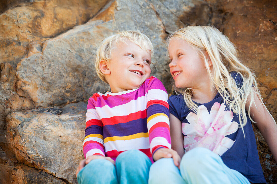 Junge und Mädchen auf Felsen, sitzen gemeinsam am Felsen und lachen, Port de Soller, Mallorca, Balearen, Spanien, Europa