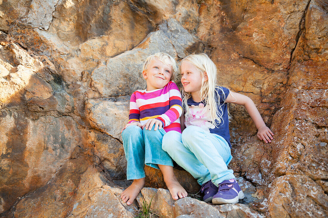 Junge und Mädchen auf Felsen, sitzen gemeinsam am Felsen und lachen, Port de Soller, Mallorca, Balearen, Spanien, Europa