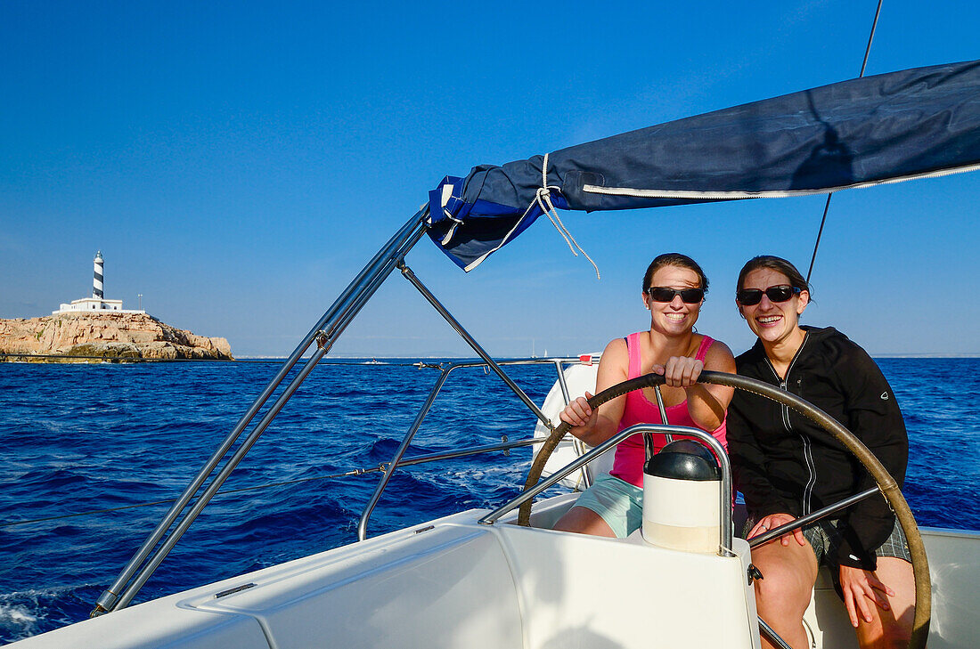 Two young women sitting behind the steering wheel of a sailing yacht steering it past the Cap Figuera with its lighthouse Faro Cala Figuera, Palma de Mallorca is still visible in the background, Balearic Islands, Spain, Europe