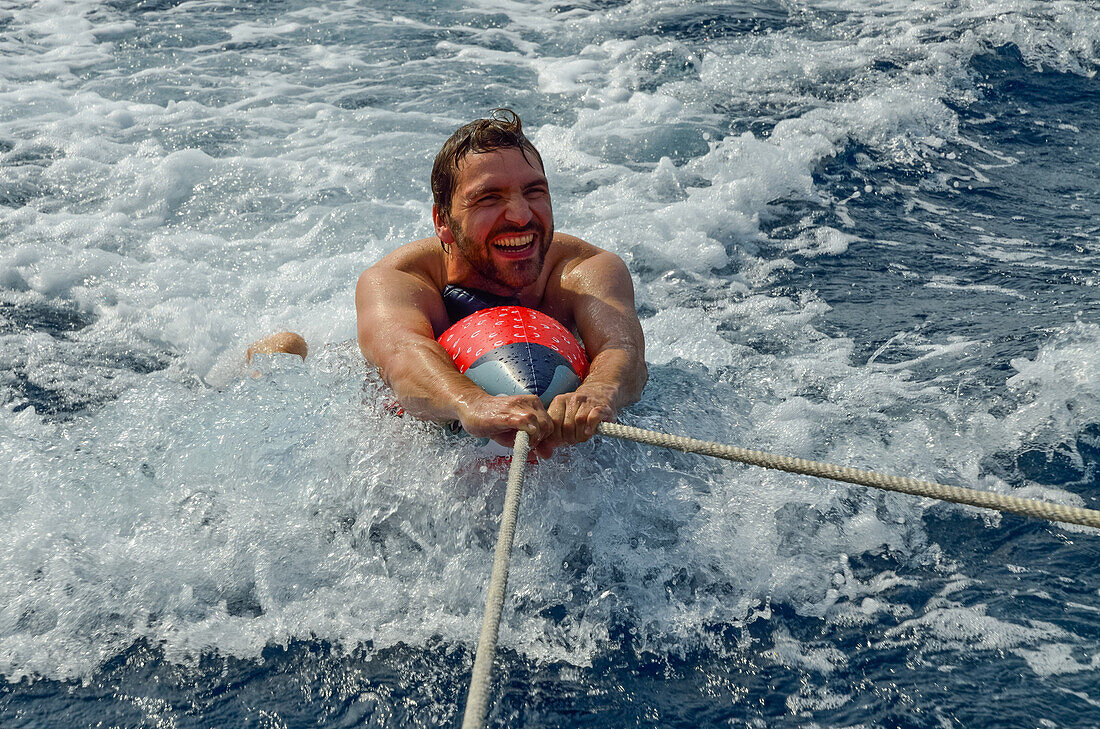Ein junger Mann wird auf einem aufblasbaren Hai hinter einer Segelyacht her gezogen und surft dabei, Mallorca, Balearen, Spanien, Europa