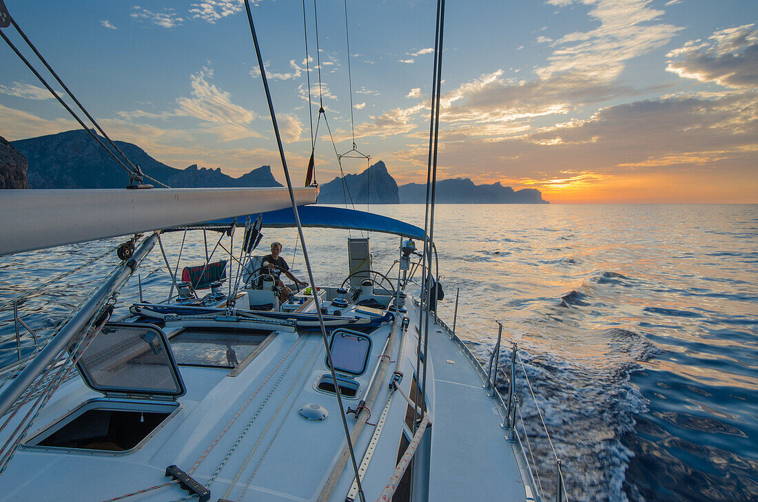 A young man sitting at the steering wheel, steering a sailing yacht at sunset, Mallorca, Balearic Islands, Spain, Europe