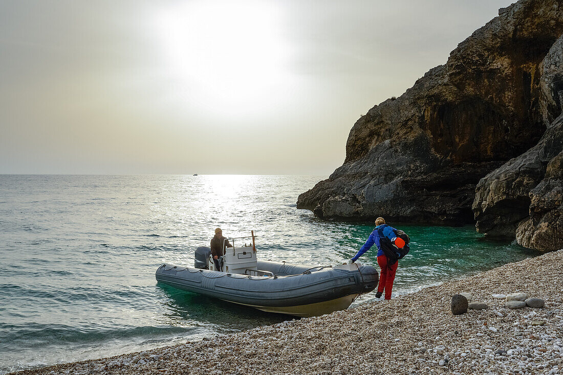 Junge Frau mit Trekkingausrüstung steigt bei Sonnenaufgang in ein Schlauchboot am Kies-Strand der Bucht Cala Sisine das sie zurück zum Ausgangspunkt des Selvaggio Blu bringt, Golfo di Orosei, Sardinien, Italien, Europa