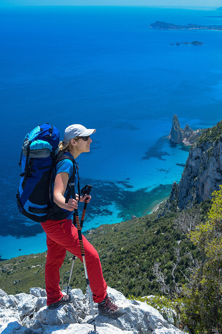 Eine junge Frau wandert entlang der gebirgigen Küste, Felsnadel bei Pedra Longa und Santa Maria Navarrese im Hintergrund, Punta Giradili, Selvaggio Blu, Sardinien, Italien, Europa