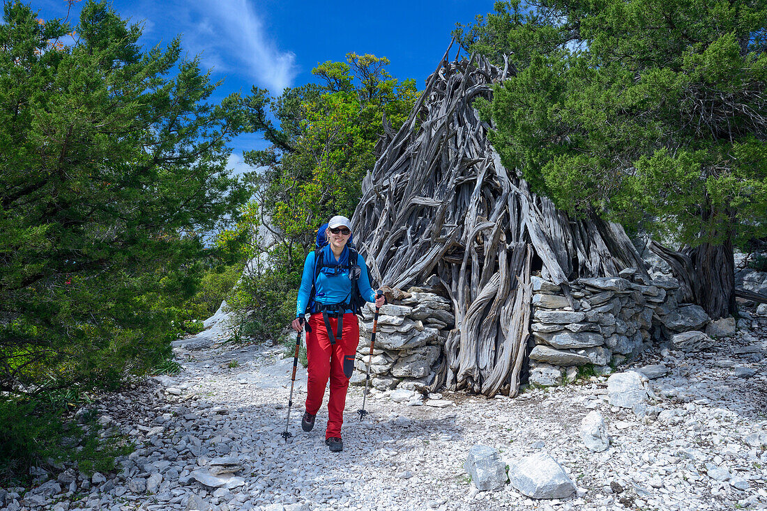 A young woman with trekking gear hiking through the mountainous coastal landscape, passing an Ovile, which is a traditional shepherds hut, Selvaggio Blu, Sardinia, Italy, Europe