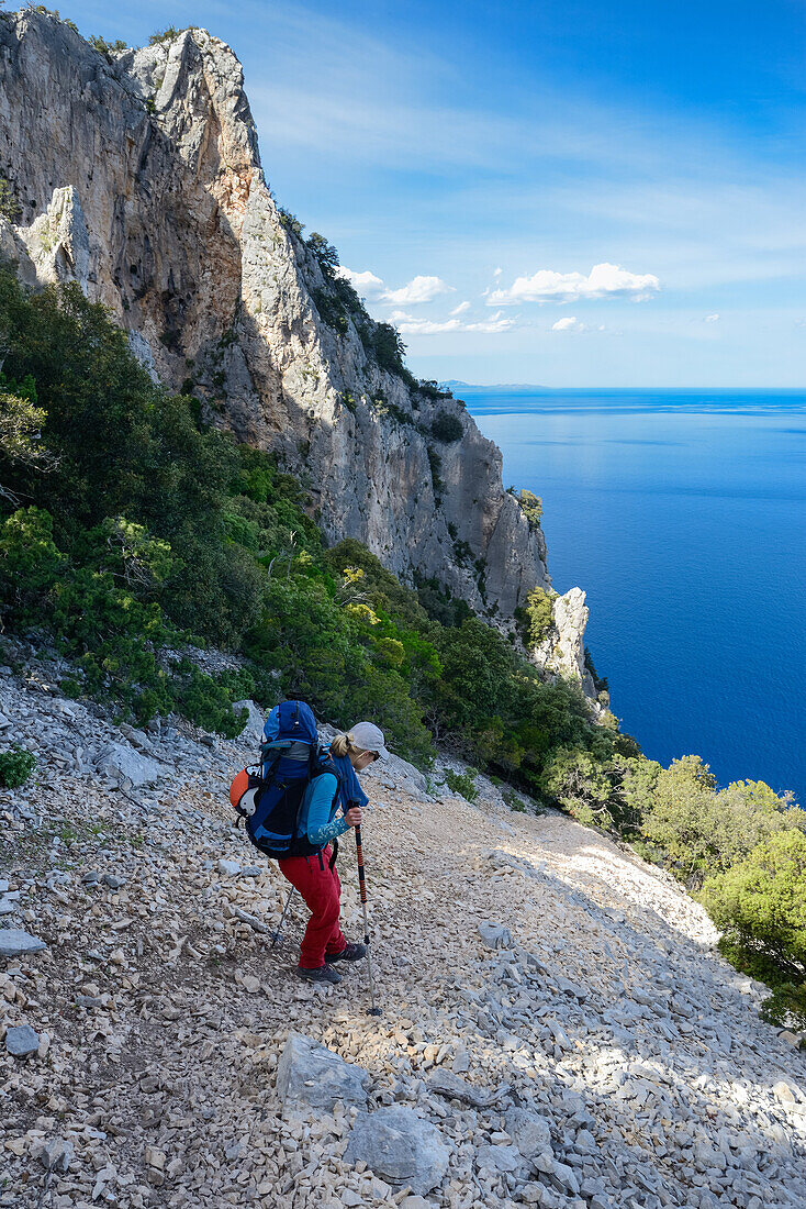 Eine junge Frau mit Trekkingausrüstung wandert oberhalb des Meeres ein steiles Geröllfeld hinunter, entlang der gebirgigen Küste, Golfo di Orosei, Selvaggio Blu, Sardinien, Italien, Europa
