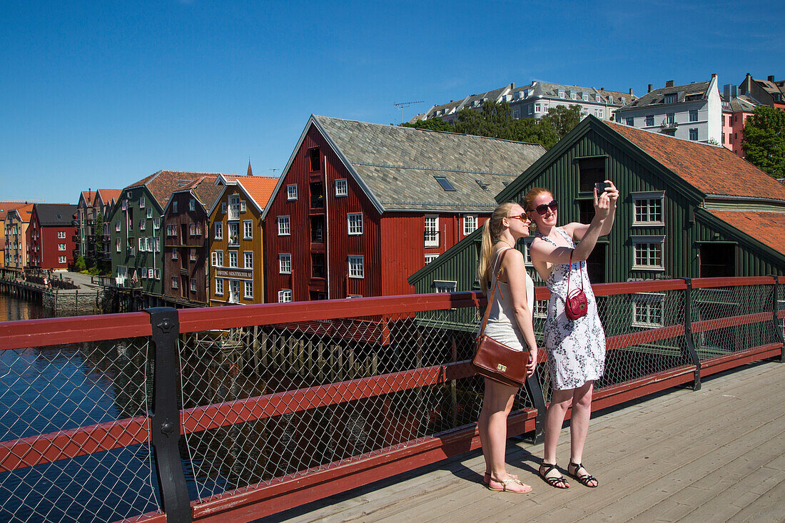 Two young girls on Gamle Bybro Old Town Bridge take selfie photograph with colorful buildings of historic wharves along river Nidelva behind, Trondheim, Sør-Trøndelag, Norway