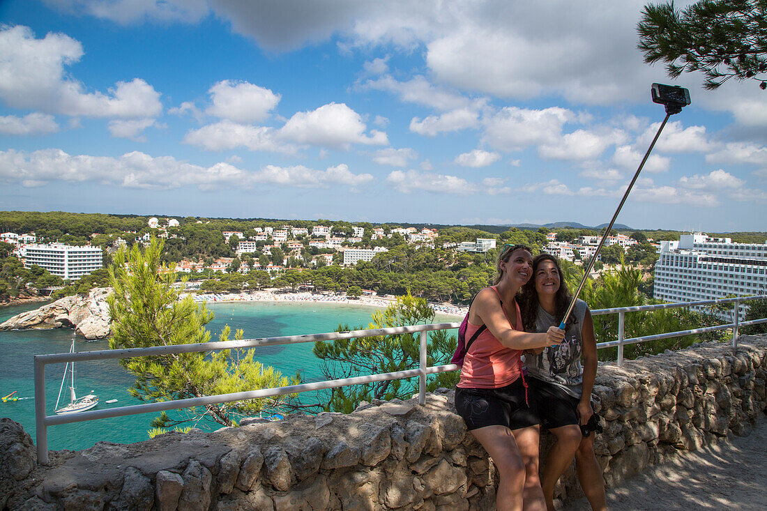 Two young women take selfie photograph overlooking Cala Galdana bay and beach with smartphone on extension stick, Cala Galdana, Menorca, Balearic Islands, Spain