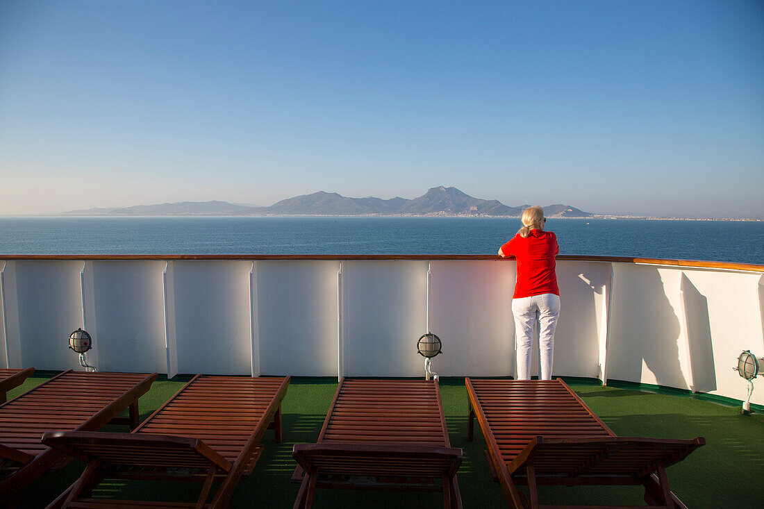Woman stands at railing of cruise ship MS Deutschland (Reederei Peter Deilmann) and looks towards Tunisian coastline, near La Goulette, Tunis, Tunisia