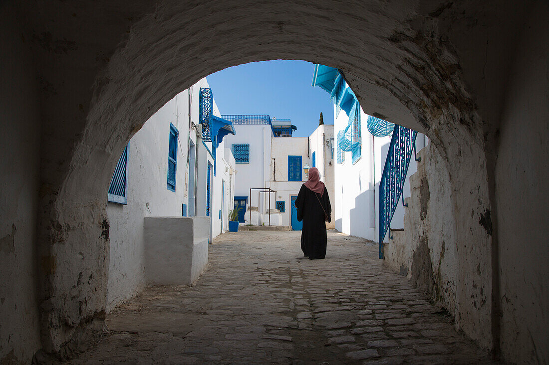 Blick durch Torbogen auf arabische Frau auf Weg entlang Häuser mit weißen Wänden und blauen Fenstern im Künstlerdorf, Sidi Bou Said, Tunis, Tunesien, Afrika