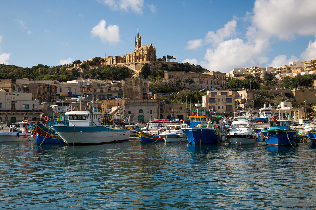 Traditionell bunt bemalte Fischerboote im Hafen und die Kapelle Unsere Liebe Frau von Lourdes auf Hügel, Mgarr, Gozo, Malta, Europa