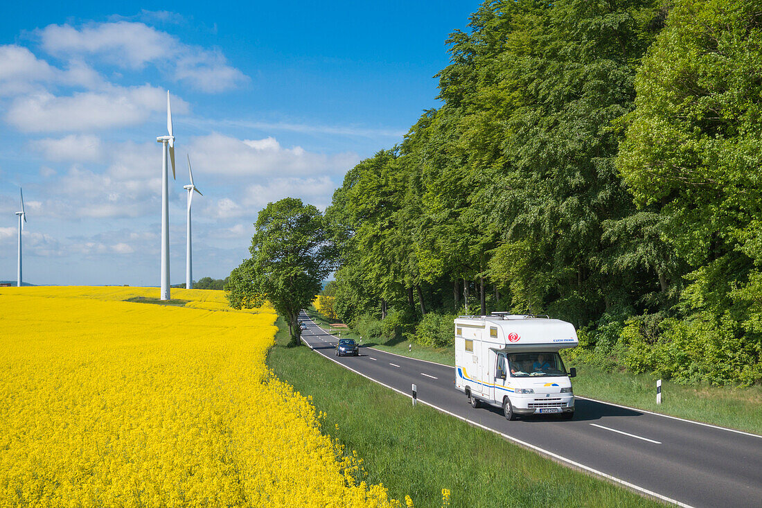 Motorhome camper on rural road and wind turbines in blooming canola field, near Alsfeld, Vogelsberg, Hesse, Germany