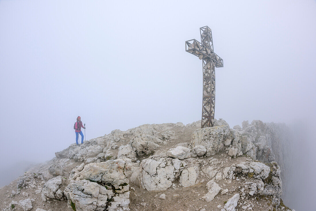 Woman hiking in fog towards cross at summit of Plattkofel, Plattkofel, Langkofel range, UNESCO world heritage Dolomites, Dolomites, Trentino, Italy