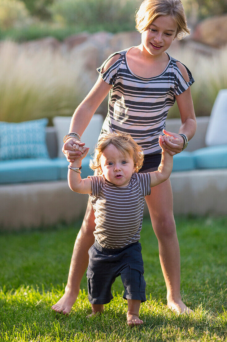 Caucasian sister helping baby brother walk in backyard, C1