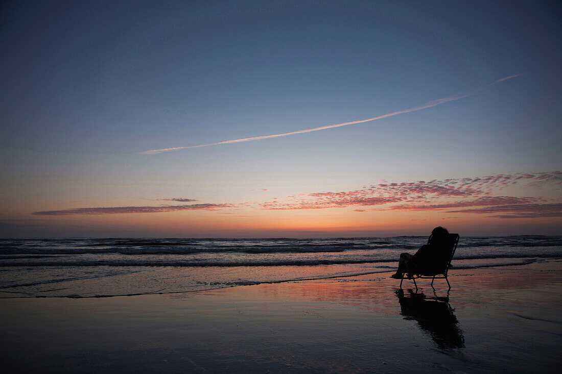 Caucasian girl in deck chair overlooking sunset on beach, C1