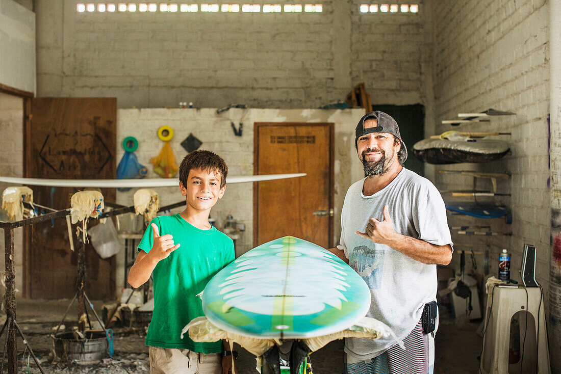Father and son gesturing hang ten in surfboard workshop, C1
