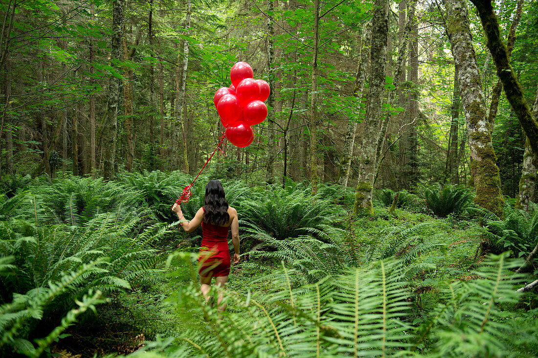 Korean woman holding red balloons in lush forest, Bainbridge Island, WA, United States