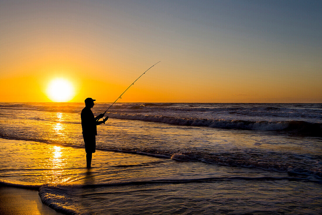 Silhouette of man fishing in waves on beach at sunset, Coast, New York, USA