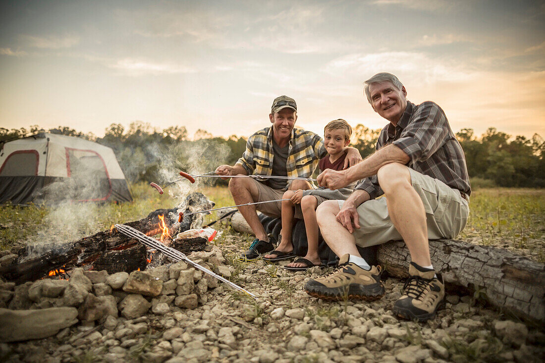 Three generations of Caucasian men roasting hot dogs over campfire, Saint Louis, Missouri, USA