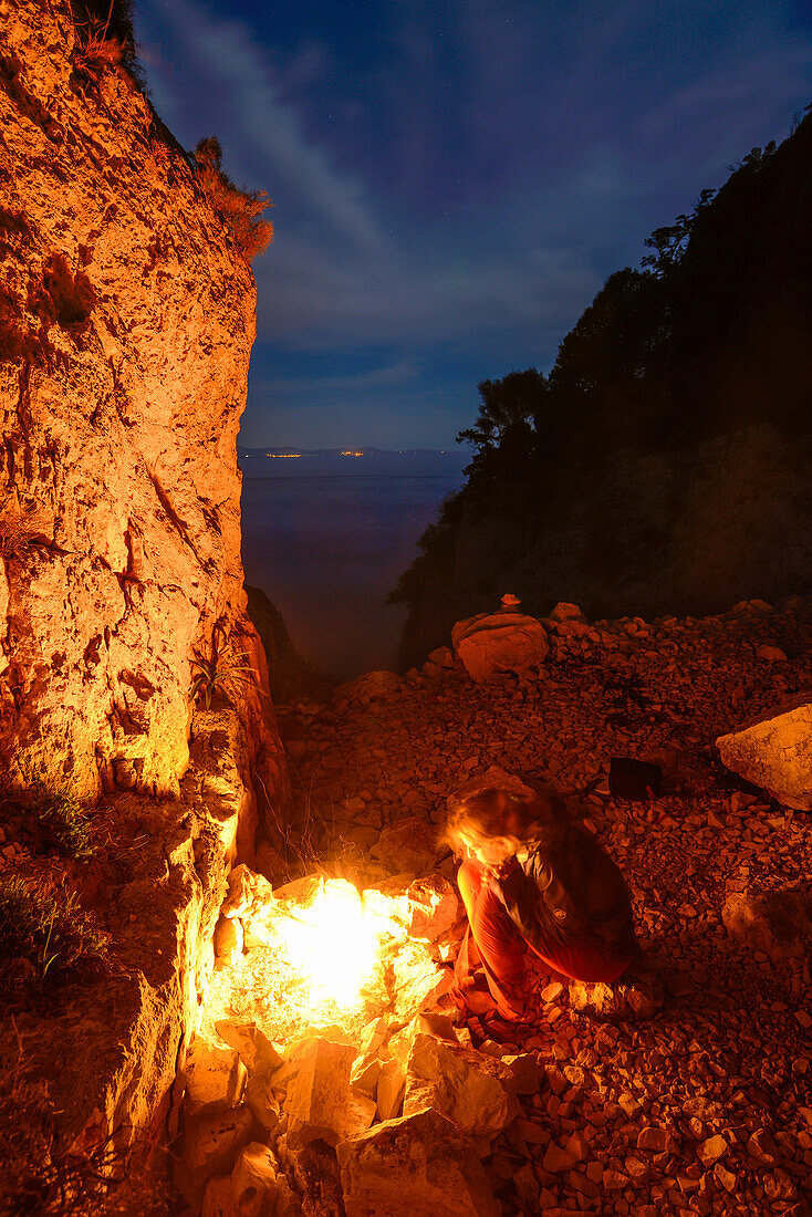 A young woman sitting at the camp fire above the sea near Bacu Mudaloru in the mountainous coast landscape, Golfo di Orosei, Selvaggio Blu, Sardinia, Italy, Europe