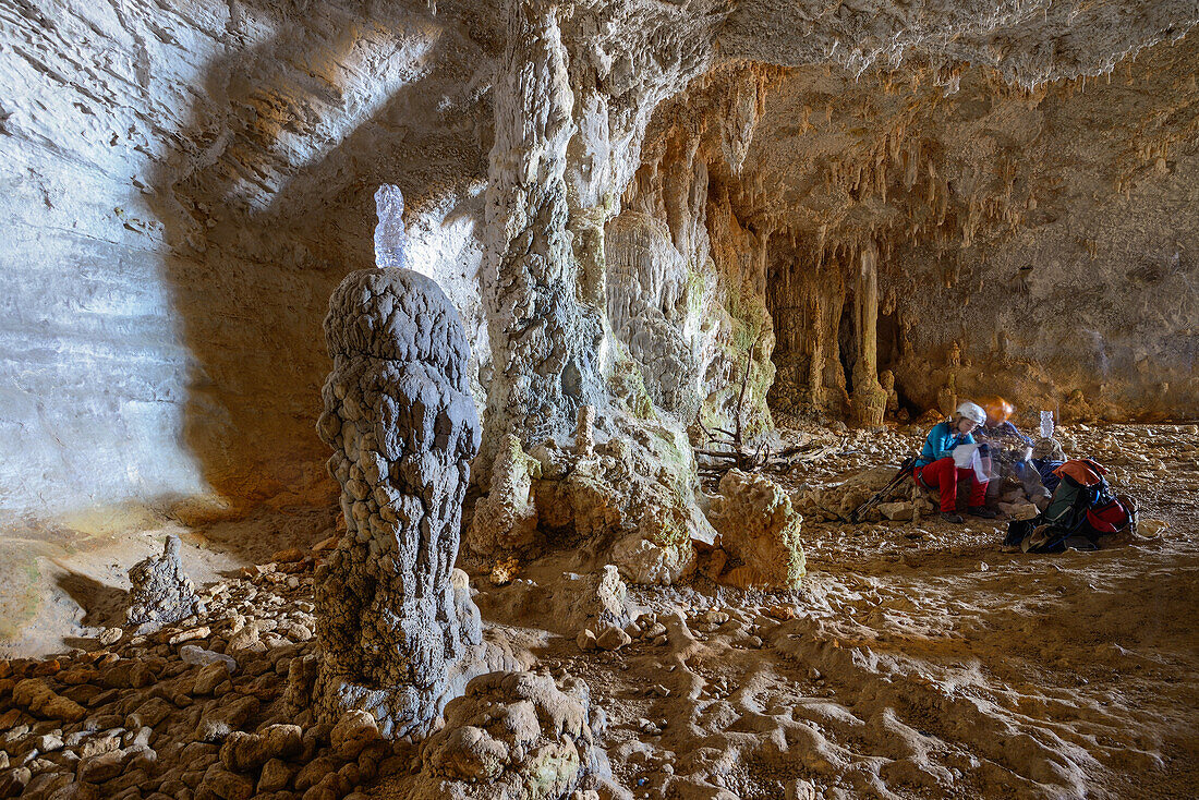 A young woman and a young man with trekking gear sitting and studying a map in a huge cave with dripstones above the sea, in mountainous coastal landscape, Golfo di Orosei, Selvaggio Blu, Sardinia, Italy, Europe