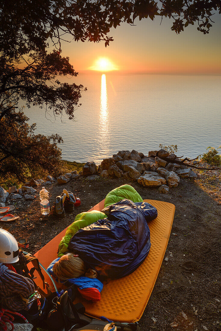 Junge Frau schläft in Schlafsack auf einem Köhlerkreis über dem Meer, bei Sonnenaufgang nahe der Bucht Cala Biriola, Golfo di Orosei, Trekking- und Kletterausrüstung, Selvaggio Blu, Sardinien, Italien, Europa