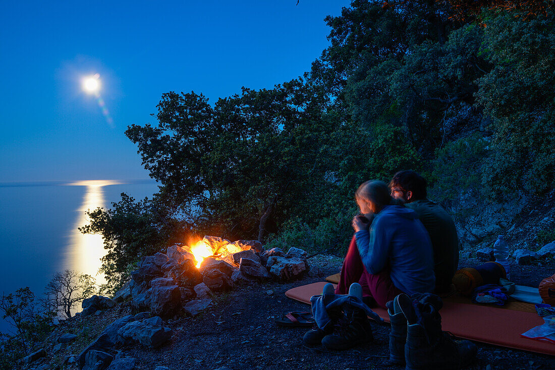 Junge Frau und junger Mann sitzen auf Isomatte am Lagerfeuer mit Vollmond auf einem Köhlerkreis über dem Meer, bei der Bucht Cala Biriola, Golfo di Orosei, Selvaggio Blu, Sardinien, Italien, Europa