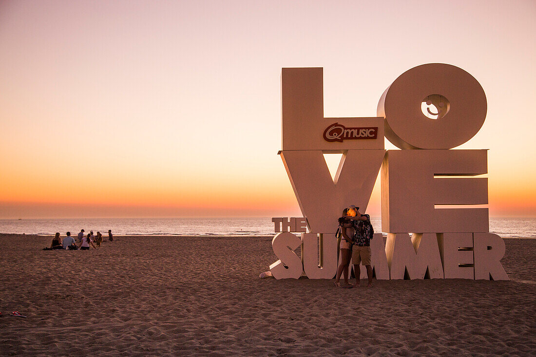 Couple taking a selfie photograph with smartphone in front of Love sculpture on the beach at sunset, Ostend, Flanders, Flemish Region, Belgium