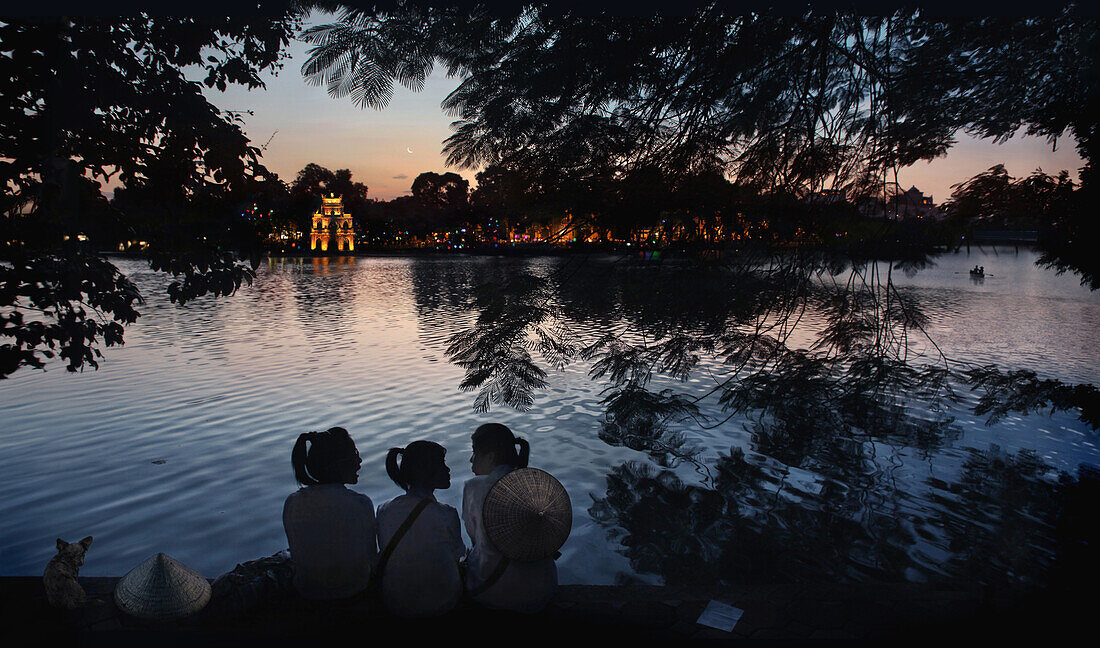 Three schoolgirls chatting on the banks of Hoan Kiem Lake with Thap Rua temple in the background, Hanoi, Vietnam, Asia