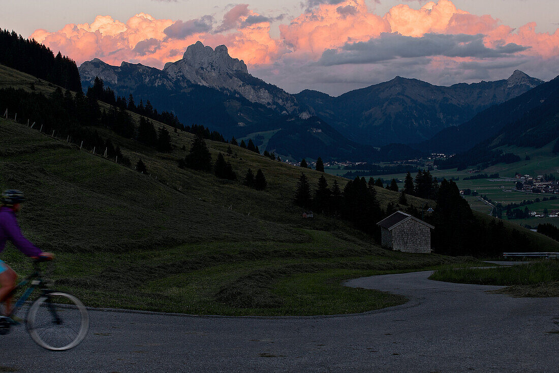 Junge Frau fährt mit ihrem Fahrad in der Nähe der Berge bei Sonnenuntergang, Rote Flüh, Gimpel, Hochwiesler, Tannheimer Tal, Tirol, Österreich